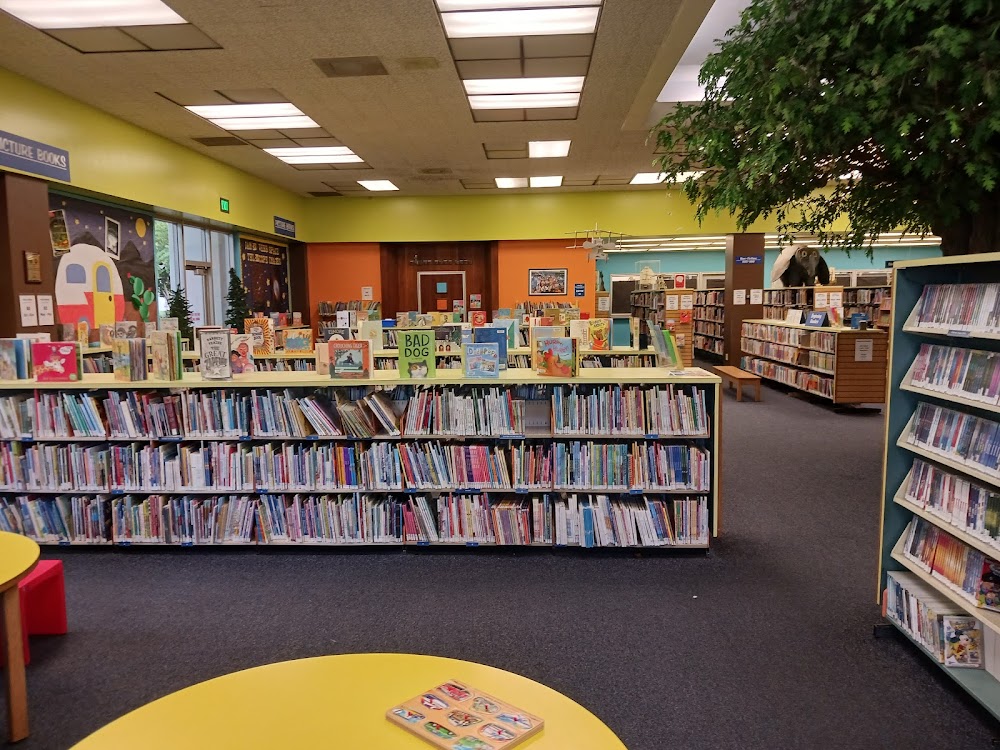 Interior view of Stanislaus County Library in Modesto, featuring colorful bookshelves filled with children's books, reading areas with round tables, and decorative elements like trees and wall art.