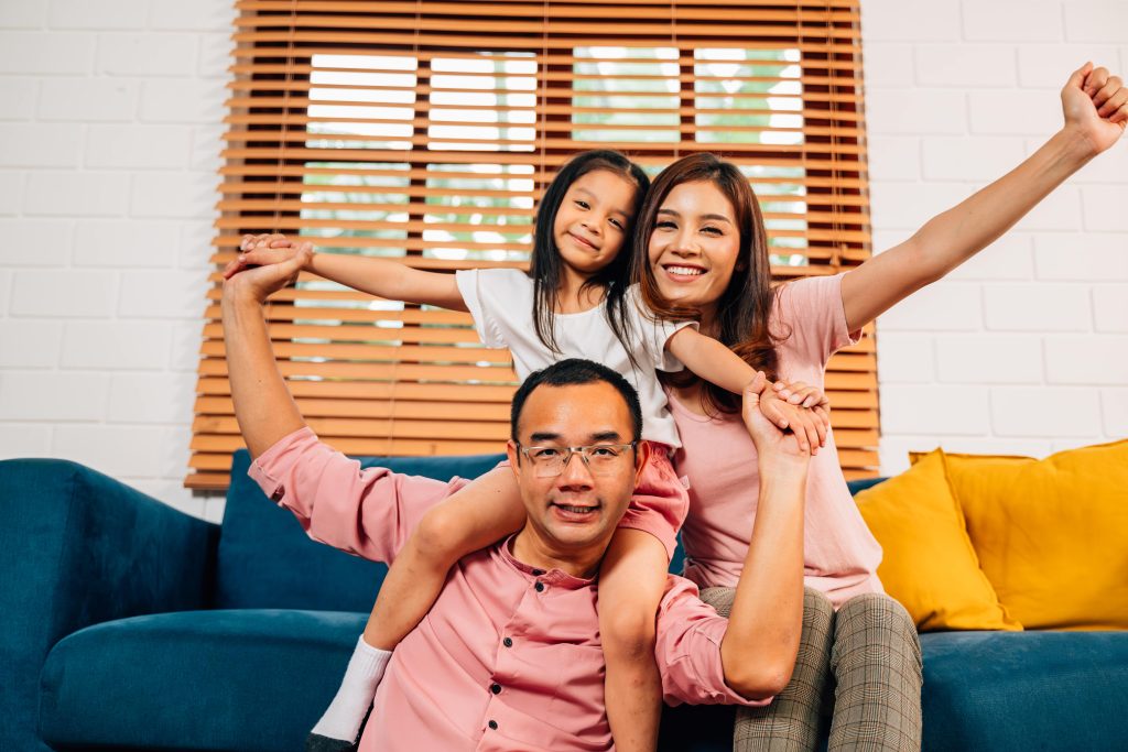 Family enjoying quality time in a living room, mother and daughter smiling, father lifting daughter on his back, bright and comfortable setting, emphasizing home comfort and family bonding.