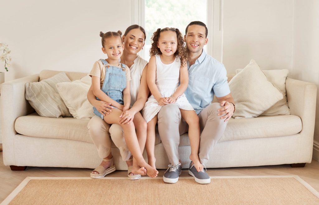 Young family of four sitting together on a beige sofa in a cozy living room, smiling and enjoying quality time, emphasizing comfort and togetherness.