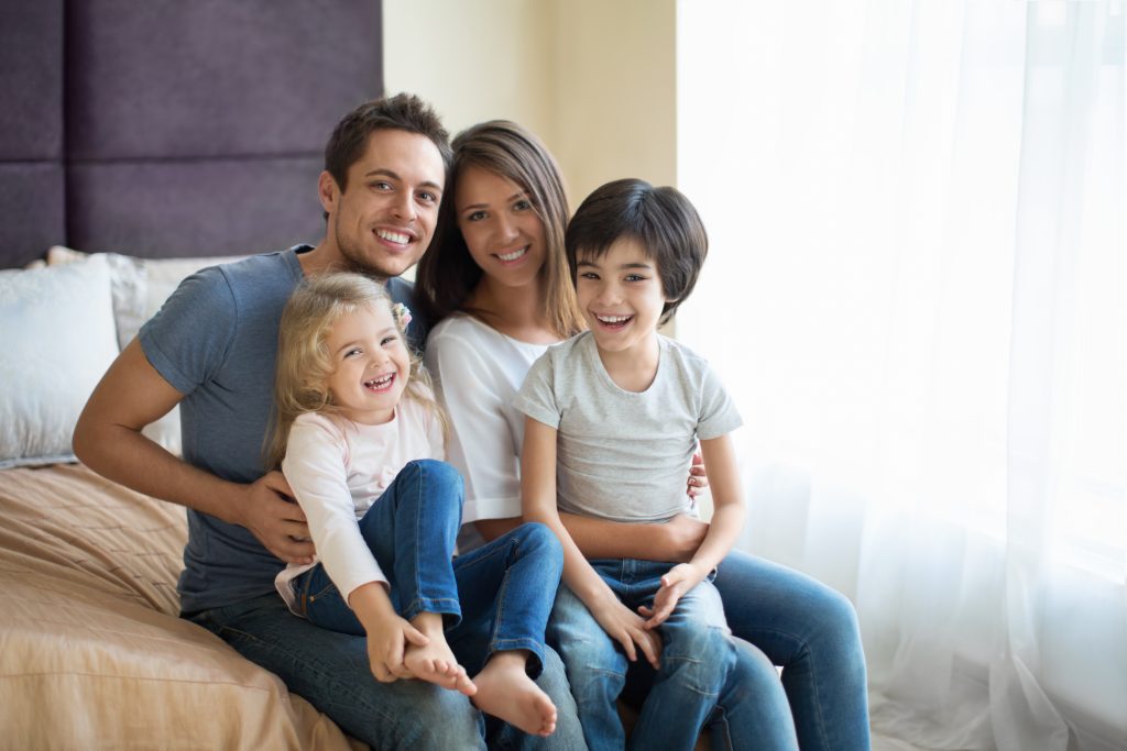 Smiling family of four sitting together on a bed, representing warmth and comfort, emphasizing the importance of a cozy home environment related to efficient furnace maintenance in Modesto, CA.