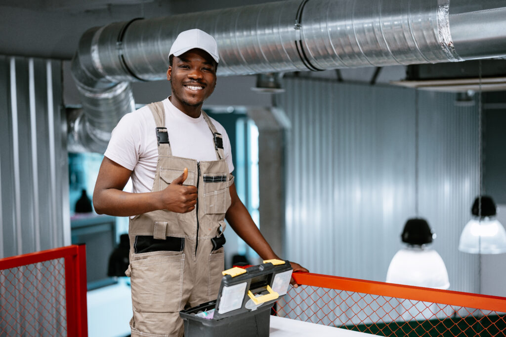 Smiling handyman in work attire giving a thumbs up, showcasing confidence in commercial HVAC maintenance services, with tools nearby in a modern workplace setting.